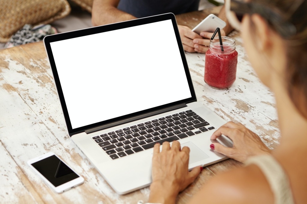 Close-up rear view of young businesswoman working on generic laptop at desk with mobile phone and sm